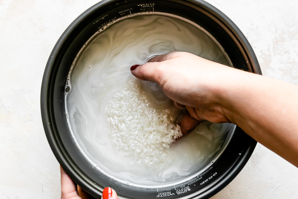 A woman's hand works to rinse white rice in water inside of the inner cooking pan of a Zojirushi rice cooker. The pan sits atop a creamy white textured surface.