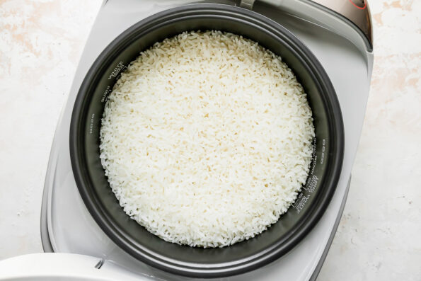 An overhead shot of perfectly cooked white rice for sushi stacks rests inside of an open Zojirushi rice cooker. The rice cooker sits atop a creamy white textured surface.