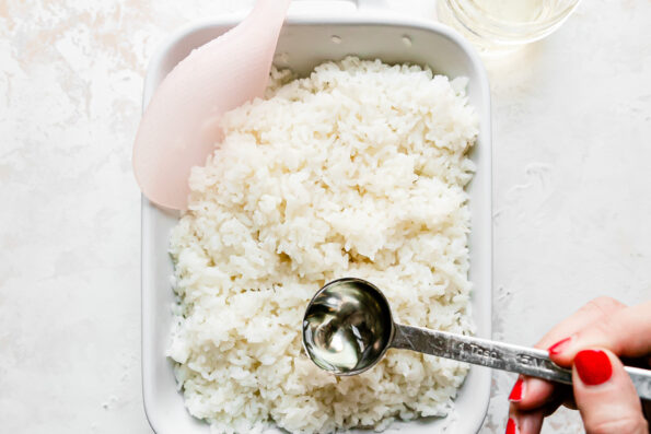Sushi rice fills a small white Staub dish that sits atop a creamy white textured surface. A woman's hand works to season the rice with a prepared sushi rice seasoning liquid using a tablespoon. A light pink rice paddle rests inside of the dish for mixing. A small glass mason jar filled with seasoning liquid rests alongside the dish.