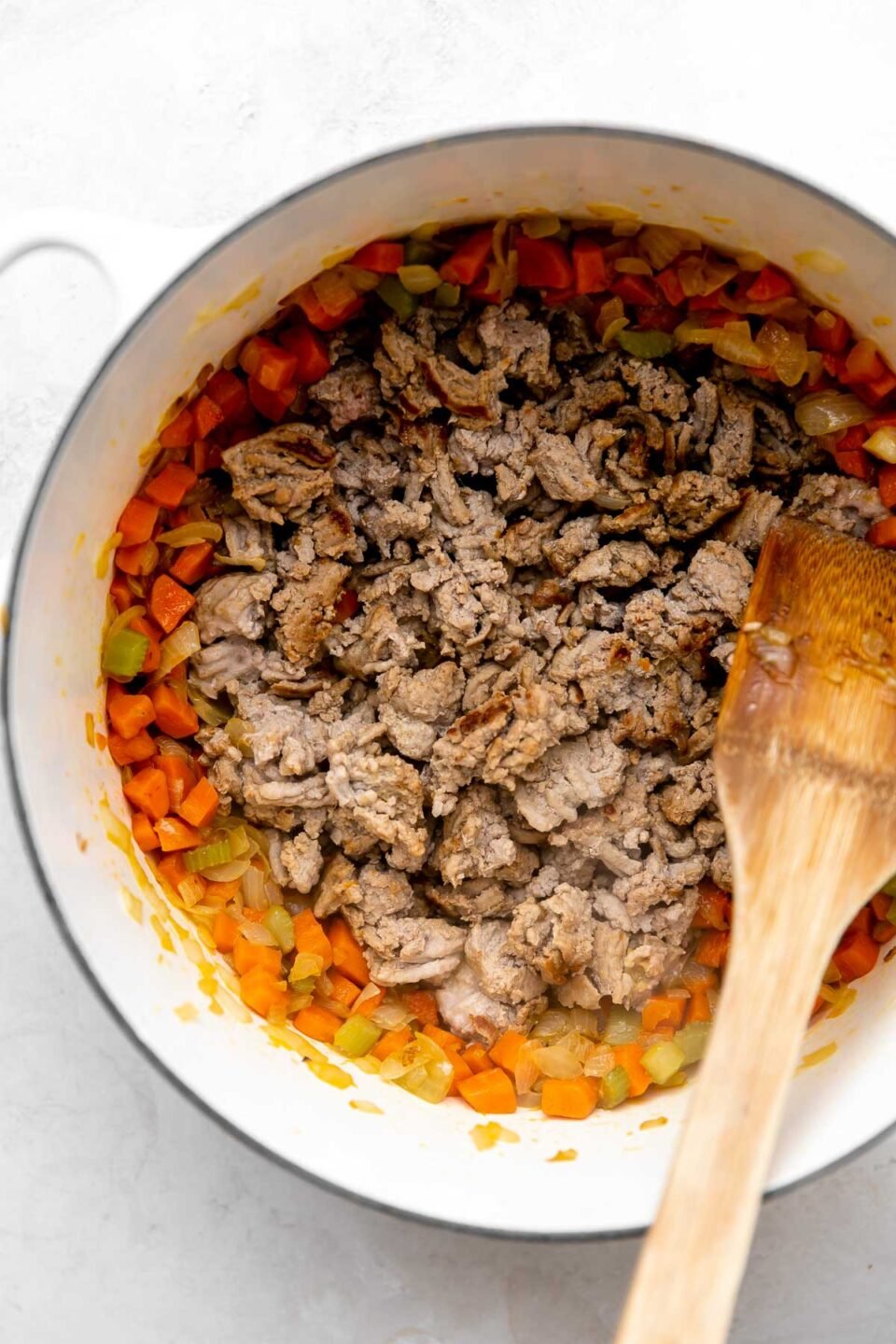 Soffrito mixture is pushed to the outer edges of a white pot, and the browned turkey sits in the center. The pot is sitting on a white and grey marbled surface. A wooden spoon rests on top of the pot.