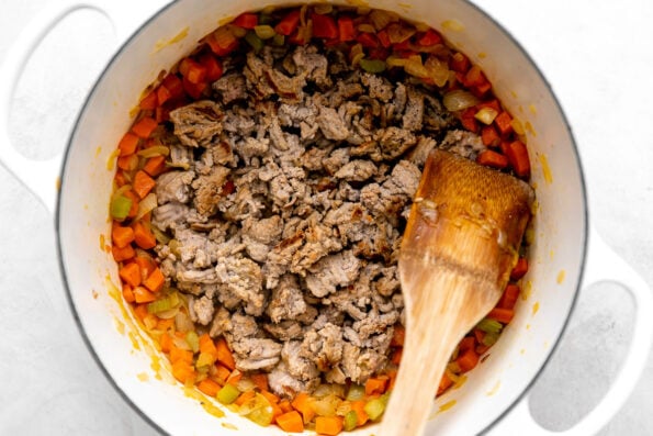 Soffrito mixture is pushed to the outer edges of a white pot, and the browned turkey sits in the center. The pot is sitting on a white and grey marbled surface. A wooden spoon rests on top of the pot.