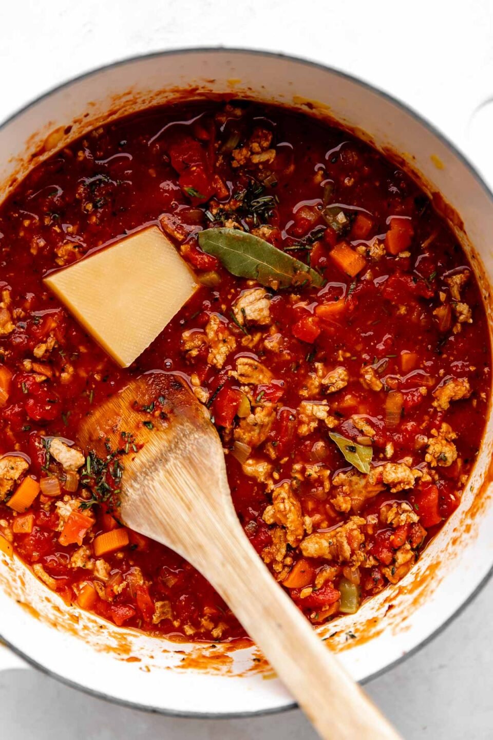 A white pot filled with bolognese sauce sits on a white and grey marbled surface. Bay leaves, fresh herbs and a parmesan rind are sitting on top of the sauce, and a wooden spoon rests in the center of the skillet.