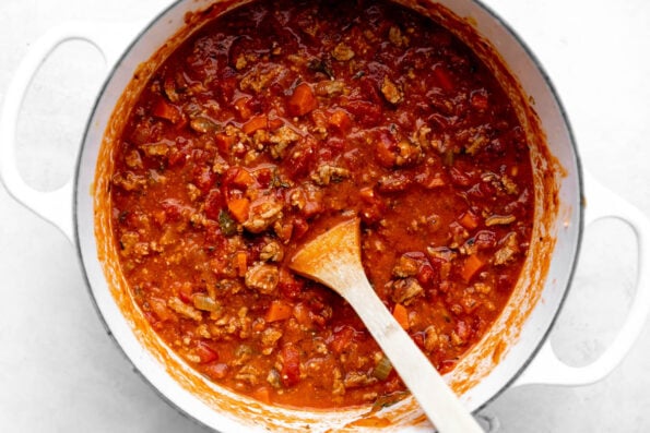 An overhead shot of turkey bolognese sauce simmering in a white handled pot on a white surface, with a wooden spoon resting in it.