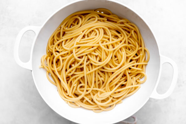Strained bucatini pasta rests in a white colander atop a white and grey marbled surface.
