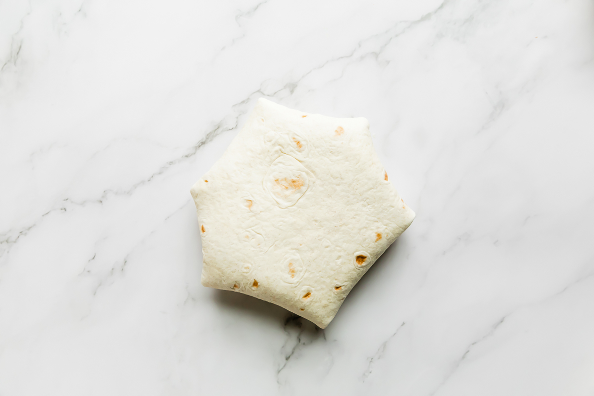 An assembled buffalo chicken crunchwrap rests seam side facing down atop a white and gray marble surface.
