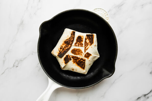 An assembled and toasted buffalo chicken crunchwrap rests inside of a white cast iron skillet that sits atop a white and gray marble surface.