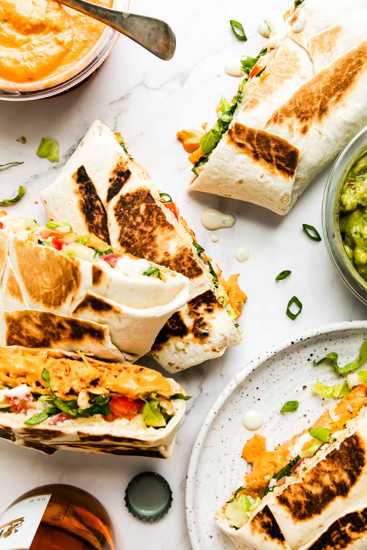 Five buffalo chicken crunchwraps are arranged atop a white and gray marble surface. An open container of Good Foods Plant Based Buffalo Dip, a second open container of Good Foods Chunky Guacamole, and a bottle of beer rest alongside the crunchwraps.