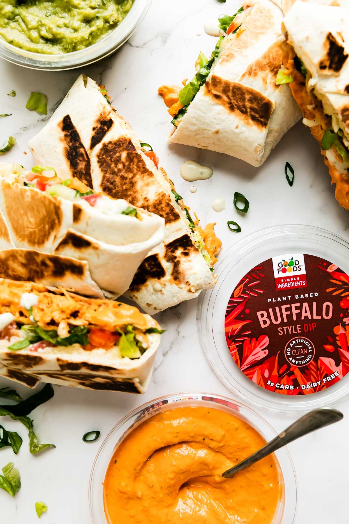 Five buffalo chicken crunch wraps are arranged atop a white and gray marble surface. An open container of Good Foods Plant Based Buffalo Dip and a second open container of Good Foods Chunky Guacamole rest alongside the crunchwraps. A spoon rests inside of the Good Foods Plant Based Buffalo Dip.