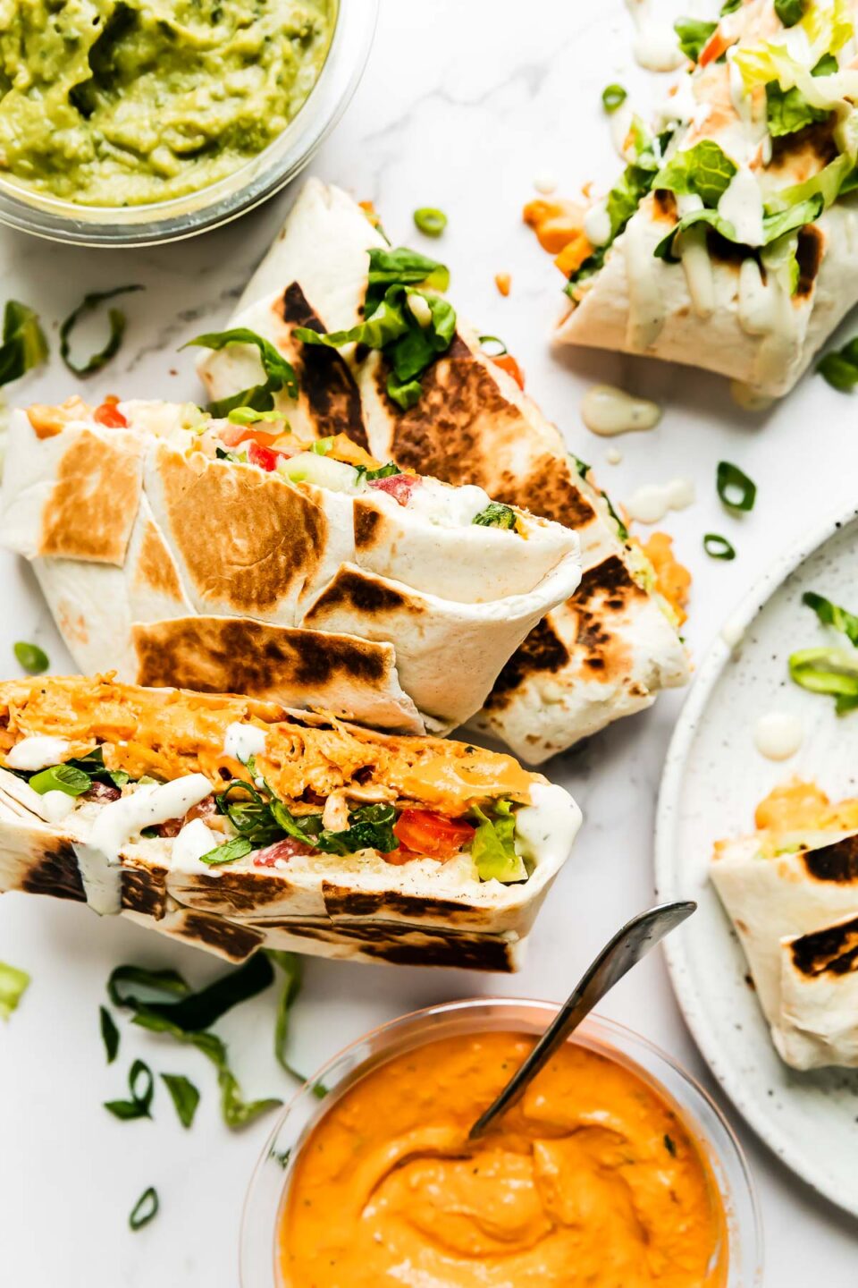 Five buffalo chicken crunch wraps are arranged atop a white and gray marble surface. An open container of Good Foods Plant Based Buffalo Dip and a second open container of Good Foods Chunky Guacamole rest alongside the crunchwraps. A spoon rests inside of the Good Foods Plant Based Buffalo Dip.
