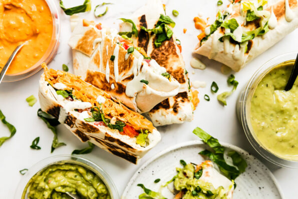 Four buffalo chicken crunchwraps are arranged atop a white and gray marble surface. An open container of Good Foods Plant Based Buffalo Dip, a second open container of Good Foods Chunky Guacamole, and a third open container of Good Foods Avocado Salsa rests alongside the crunch wraps. A spoon rests inside of the Good Foods Plant Based Buffalo Dip.