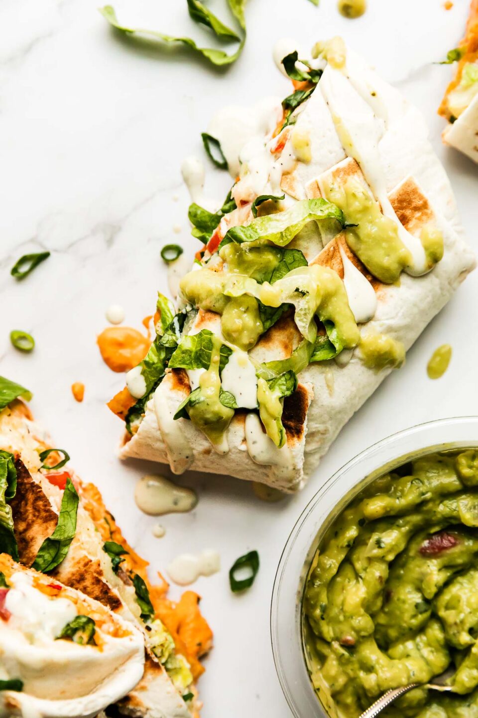 Four buffalo chicken crunchwraps are arranged atop a white and gray marble surface. An open container of Good Foods Chunky Guacamole rests alongside the crunchwraps. A spoon rests inside of the Good Foods Chunky Guacamole. The buffalo chicken crunchwrap halve at center is topped with drizzles of ranch dressing, Good Foods Avocado Salsa, and shredded lettuce.