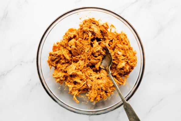 How to make a homemade crunchwrap, step 3: Shred & mix the buffalo chicken. Shredded buffalo chicken mixed with Good Foods Plant Based Buffalo Dip fills a large glass mixing bowl with a spoon resting inside the bowl. The bowl sits atop a white and gray marble surface.