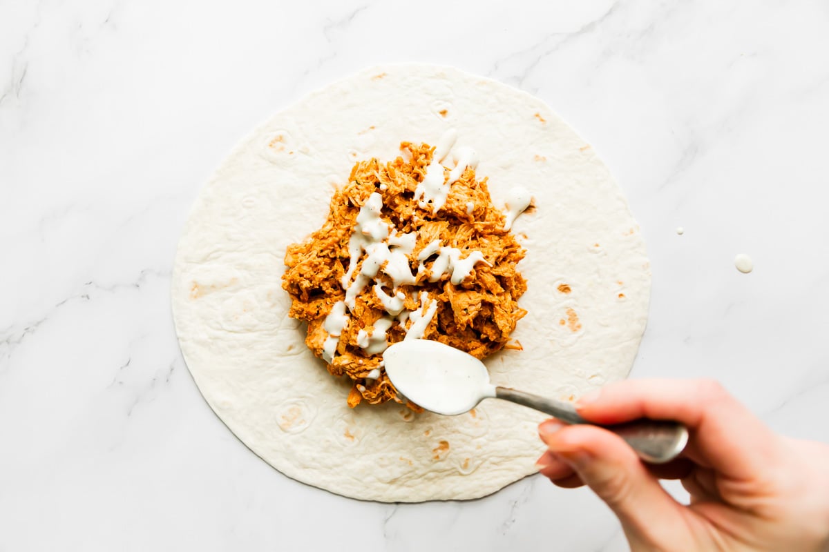 How to make a buffalo crunch wrap, step 4: Assemble the buffalo chicken crunchwraps. A flour tortilla lays flat on a white and gray marble surface. Shredded buffalo chicken has been piled atop the tortilla and a woman's hand drizzles ranch dressing using a spoon over the buffalo chicken.
