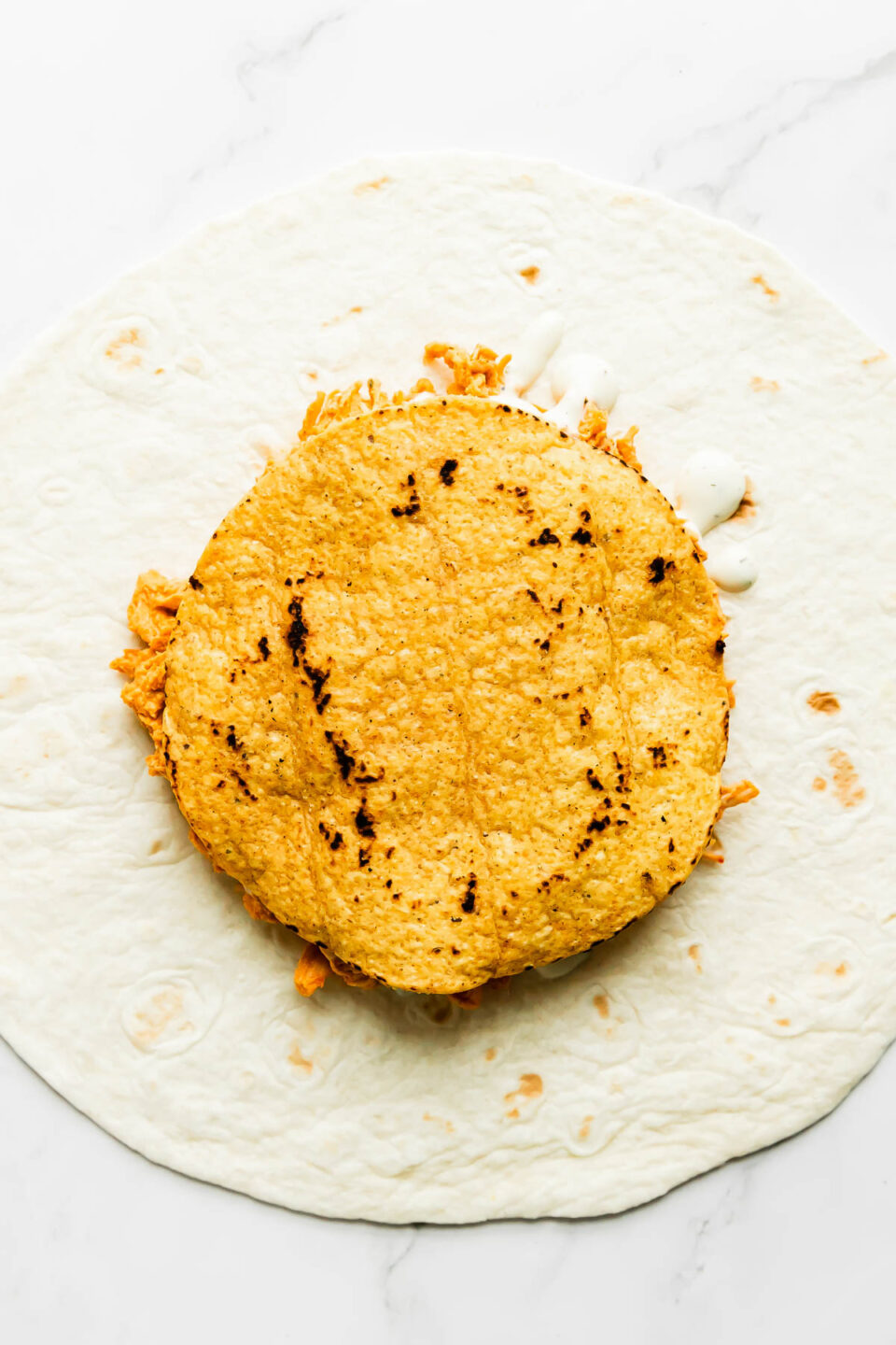 How to make buffalo crunch wraps, step 4: Assemble the buffalo chicken crunchwraps. A flour tortilla lays flat on a white and gray marble surface. A single tostada has been placed atop of shredded buffalo chicken drizzled with ranch dressing that has been piled atop the flour tortilla.