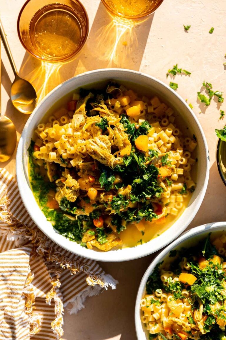 Golden soup served inside of two white ceramic bowls that sit atop a creamy peach colored textured surface. The bowls of soup are surrounded by a white and yellow striped linen napkin, amber colored drinking glasses and gold spoons.