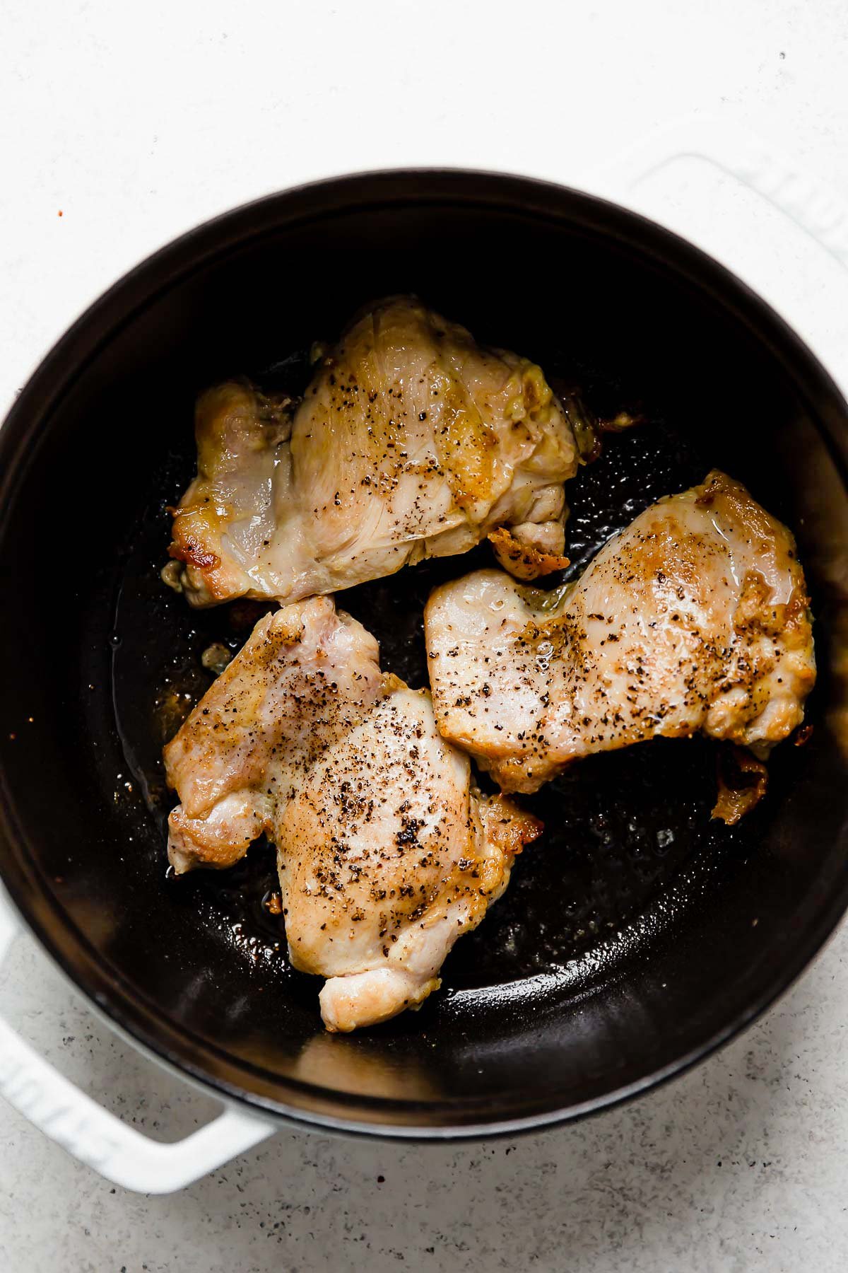 Browning boneless, skinless chicken thighs in a large white Staub cocotte for golden soup. The cocotte pot sits atop a white textured surface.