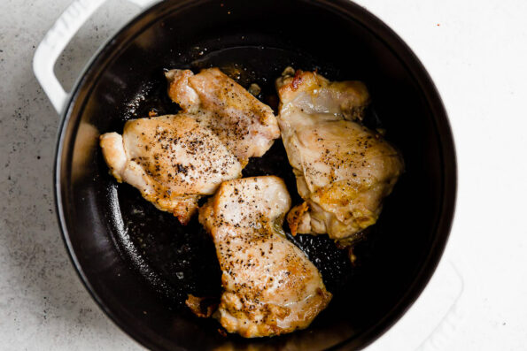 How to make golden soup, step 1: Brown the chicken. Browned chicken thighs rest inside of a white Staub cocotte that sits atop a white textured surface.