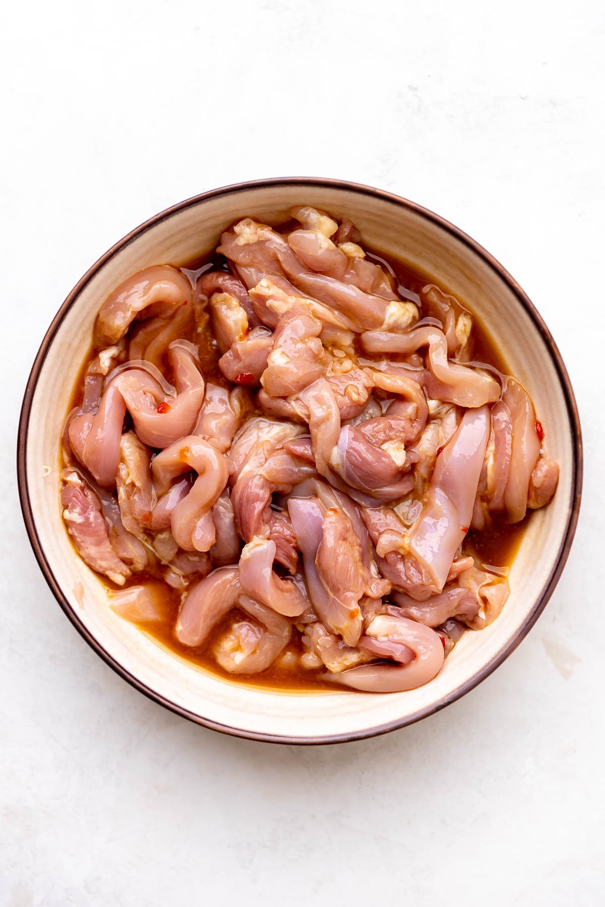 Thinly sliced chicken thighs marinate in a stir fry orange chicken marinade inside of a large ceramic bowl. The bowl sits atop a creamy white textured surface.