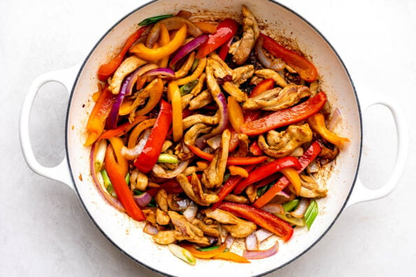 Orange chicken and vegetable stir fry fills a large white double handle skillet that sits atop a creamy white textured surface.