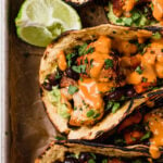A close up shot of sweet potato and cauliflower tacos drizzled with chipotle cashew crema and fresh chopped cilantro arranged atop a parchment lined baking sheet pan. The tacos are surrounded by lime halves and the baking sheet sits atop a creamy white textured surface.