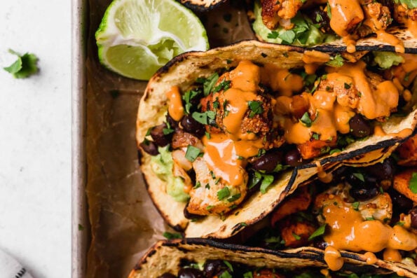 A close up shot of sweet potato tacos with cauliflower drizzled with chipotle cashew crema and fresh chopped cilantro arranged atop a parchment lined baking sheet pan. The tacos are surrounded by lime halves and the baking sheet sits atop a creamy white textured surface.