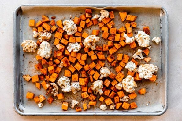 How to make cauliflower and sweet potato tacos, step 1: season the cauliflower and sweet potatoes. Cauliflower florets and diced sweet potato drizzled with olive oil and seasoned with spices are arranged atop a parchment lined baking sheet. The baking sheet sits atop a light peach colored textured surface.