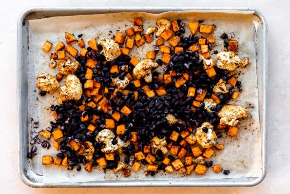 How to make roasted veggie tacos, step 2: Roast the cauliflower and sweet potatoes. Seasoned and roasted cauliflower florets and diced sweet potato are arranged atop a parchment lined baking sheet. Drained black beans are added to the sheet pan and the baking sheet rests atop a light peach colored textured surface.