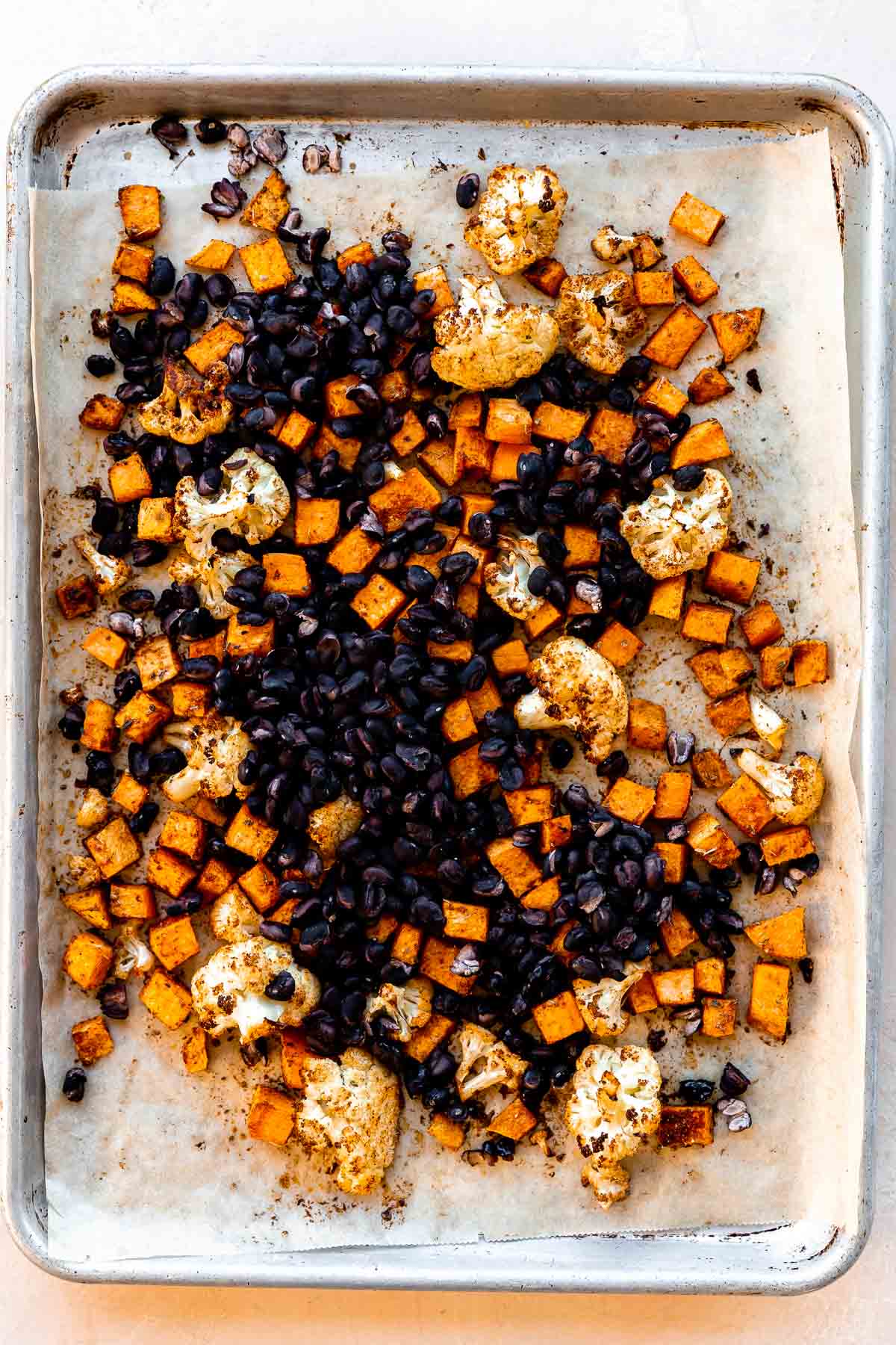 How to make roasted veggie tacos, step 2: Roast the cauliflower and sweet potatoes. Seasoned and roasted cauliflower florets and diced sweet potato are arranged atop a parchment lined baking sheet. Drained black beans are added to the sheet pan and the baking sheet rests atop a light peach colored textured surface.