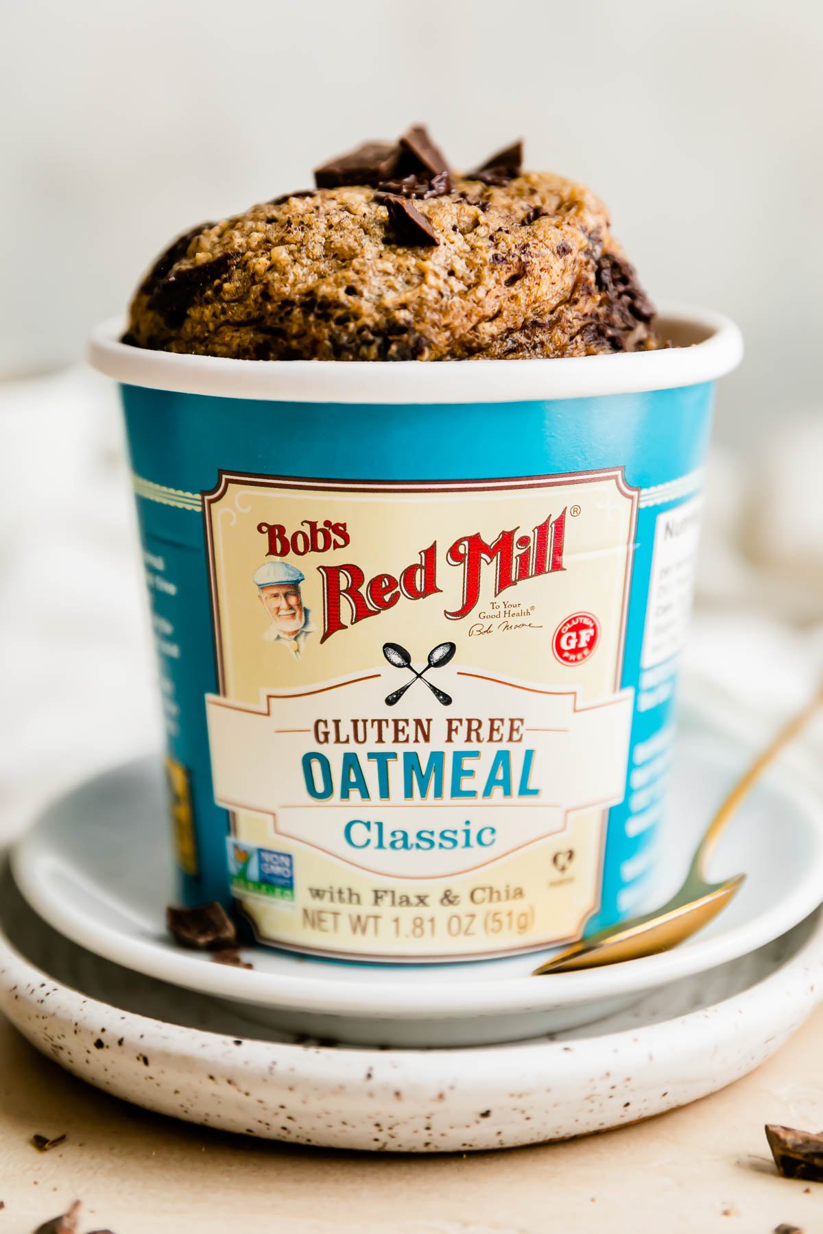 A banana oatmeal mug cake fills a Bob's Red Mill Gluten Free Classic Oatmeal Cup with Flax & Chia. The oatmeal cup sits atop two plates atop a creamy white textured surface with the label facing outwards and is surrounded by a white linen napkin. A gold spoon rests atop the plates.