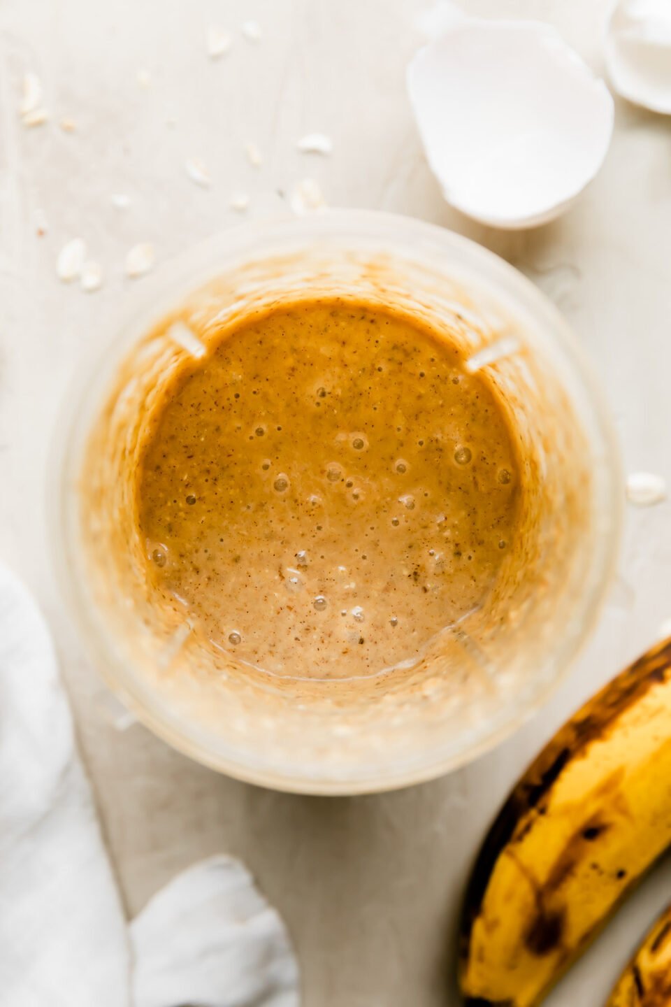 Banana oatmeal mug cake batter fills a personal blender jar that sits atop a creamy white textured surface. The jar is surrounded by a white linen napkin, loose oats, two ripe bananas, and a discarded egg shell broken in half.