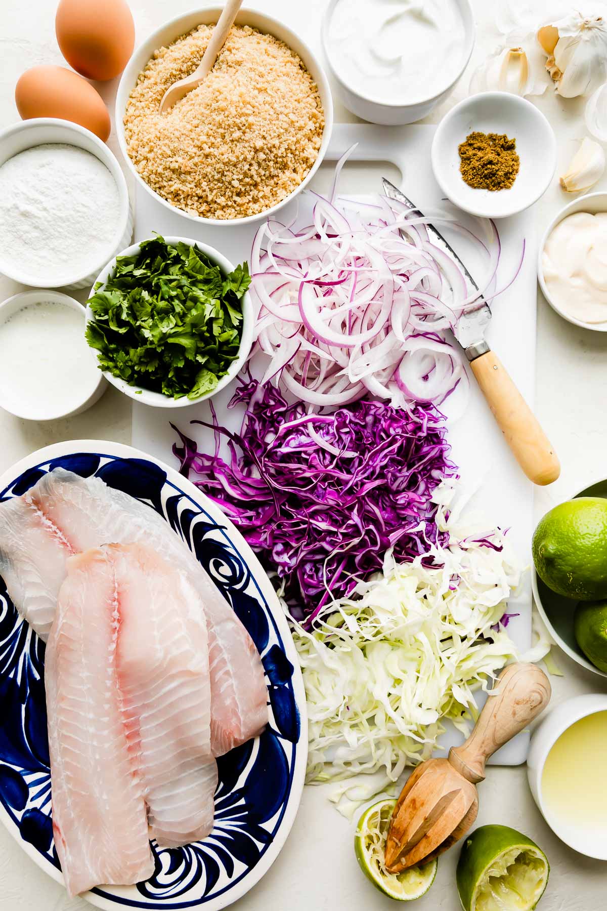 Crispy fish tacos ingredients arranged on a white textured surface: flounder, rice flour, eggs, panko breadcrumbs, cabbage, cilantro, red onion, sour cream, buttermilk, mayonnaise, garlic, lime, and ground cumin.