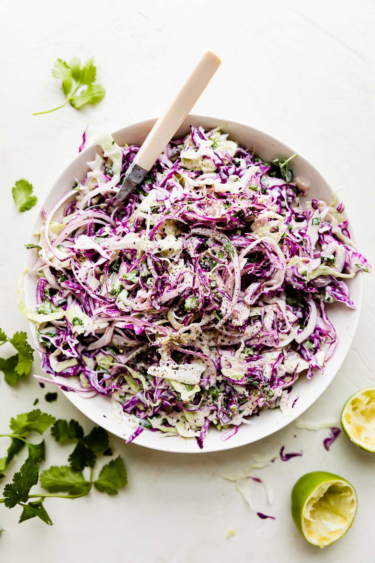 Cilantro ranch slaw fills a large white mixing bowl. The bowl sits atop a creamy white textured surface. A fork rests inside of the bowl and loose fresh herbs and spent lime halves surround the bowl at center.