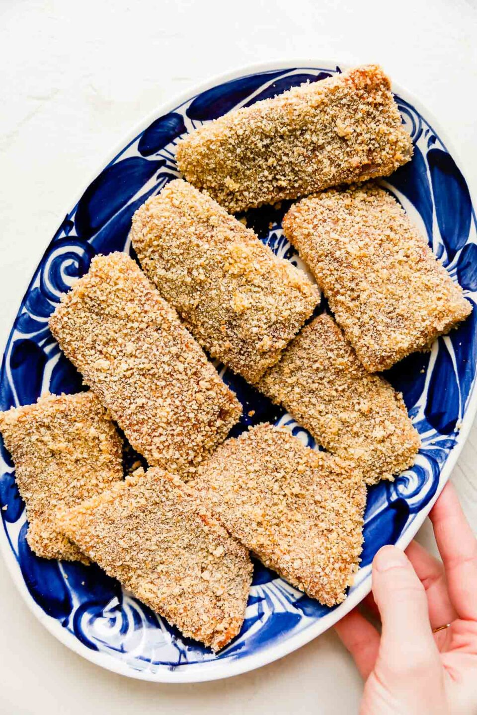 Eight panko crusted white fish fillets are arranged atop a white and blue painted platter. The platter sits atop a creamy white textured surface and a woman's hand holds the platter on one side.