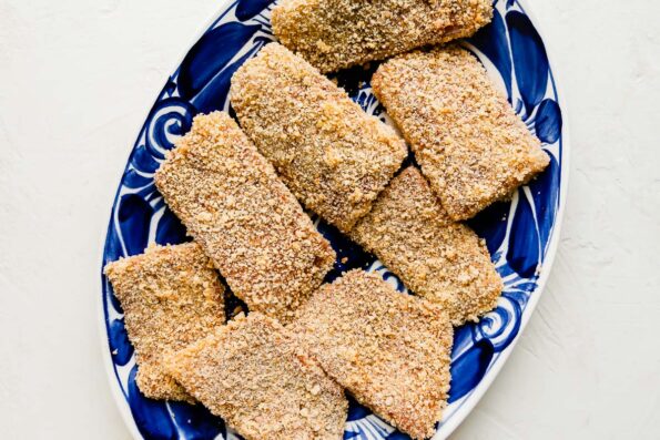 Eight panko crusted white fish fillets are arranged atop a white and blue painted platter. The platter sits atop a creamy white textured surface.