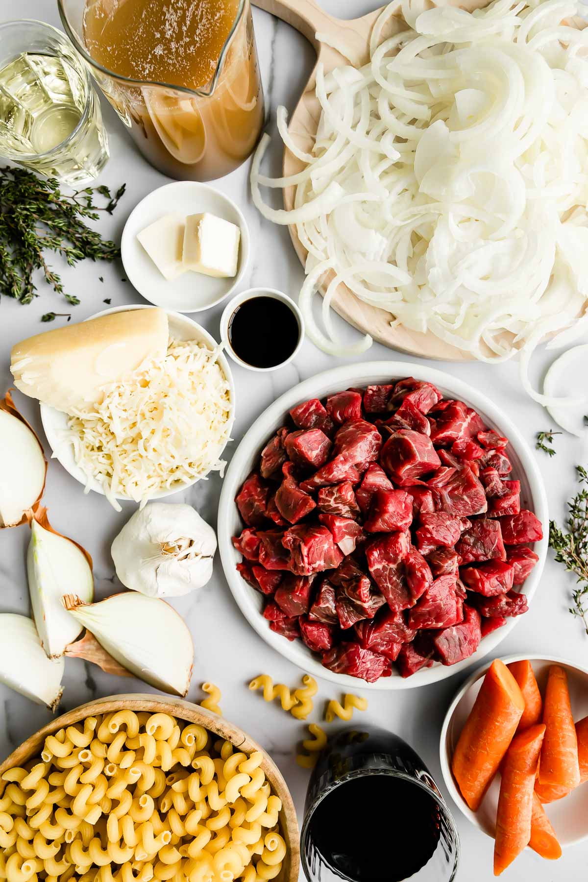 French onion pasta ingredients arranged on a white textured surface: beef stew meat, olive oil, red wine, carrots, yellow onion, garlic, fresh thyme, Worcestershire sauce, beef stock or beef broth, unsalted butter, sweet onions, sherry, cavatappi pasta, and gruyère cheese.