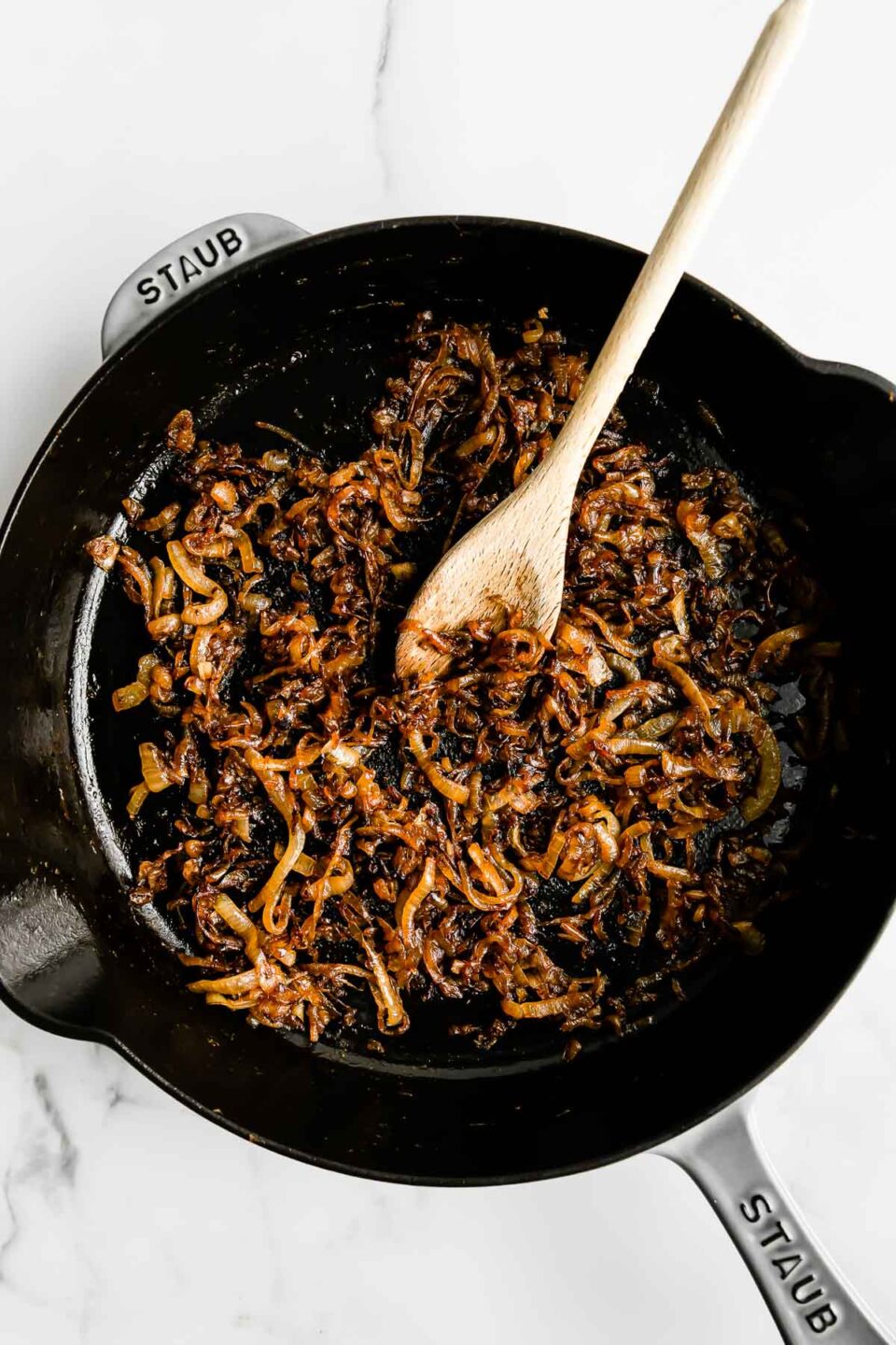 Caramelized sweet onions fill a large gray Staub cast iron skillet that sits atop a white and gray marble surface. A wooden spoon rests inside of the skillet for stirring.