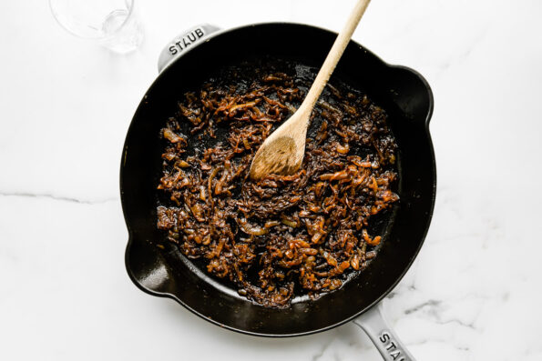 Caramelized sweet onions, deglazed with sherry fill a large gray Staub cast iron skillet that sits atop a white and gray marble surface. A wooden spoon rests inside of the skillet for stirring.