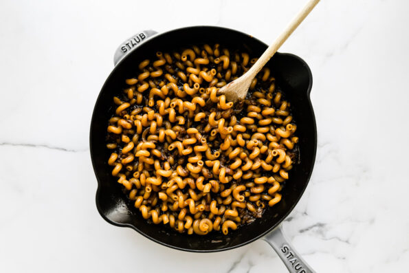 Cavatappi pasta cooks inside of reserved beef stock along with caramelized onions in a large gray Staub cast iron skillet. The skillet sits atop a white and gray marble surface and a wooden spoon rests inside of the skillet for stirring.