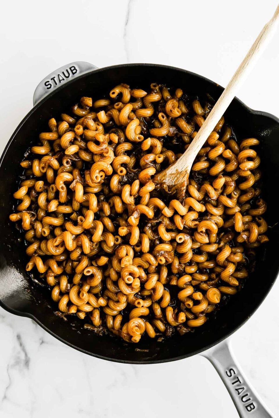 Cavatappi pasta cooks inside of reserved beef stock along with caramelized onions in a large gray Staub cast iron skillet. The skillet sits atop a white and gray marble surface and a wooden spoon rests inside of the skillet for stirring.