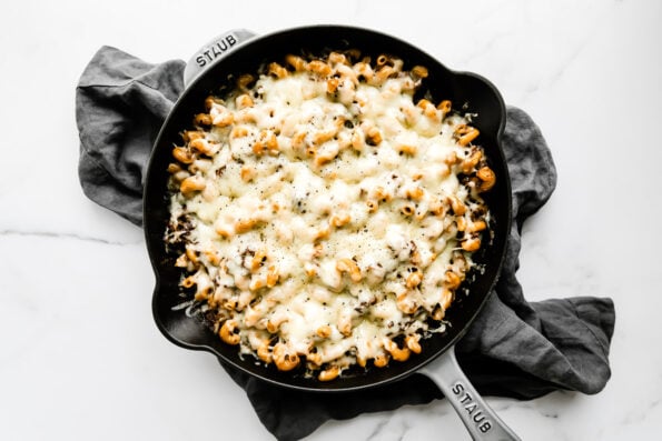 French onion pasta with braised beef and caramelized onion fills a large gray Staub cast iron skillet. The pasta has been finished with melted gruyère cheese and the skillet rests atop a white and gray marble surface with a dark gray linen napkin resting underneath the skillet at center.
