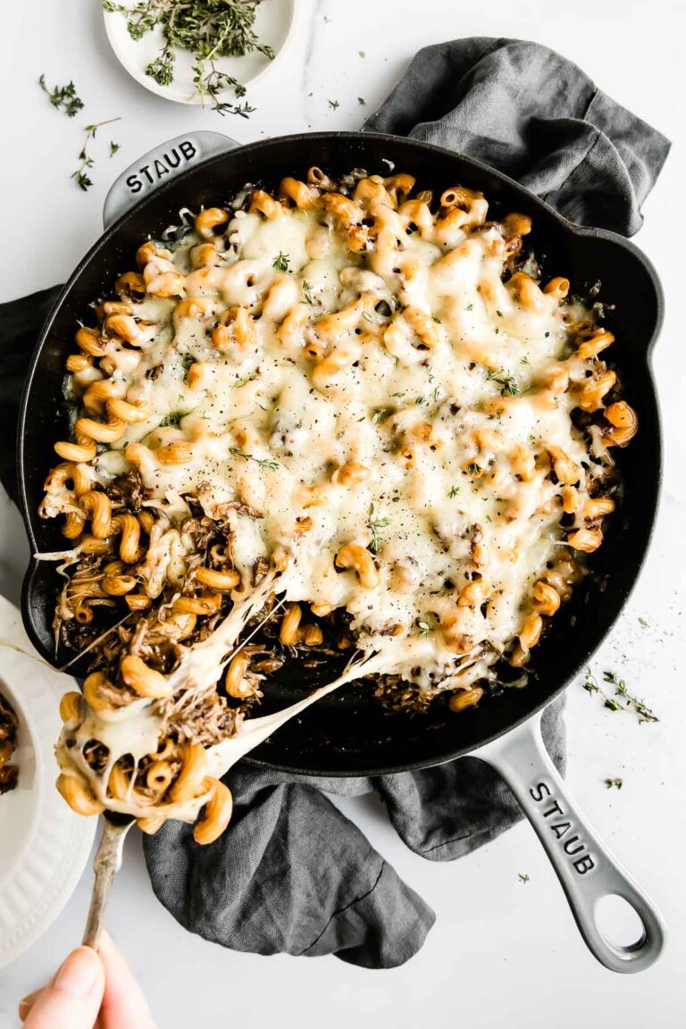 One pot french onion pasta fills a large gray Staub skillet that sits atop a white and gray marble surface. A dark gray linen napkin rests underneath the skillet and a small white bowl filled with fresh thyme rests alongside. A woman's hand uses a spoon to scoop a portion of the pasta out of the skillet and into a white bowl. A melted gruyère cheese pull is shown as the spoon leaves the skillet.