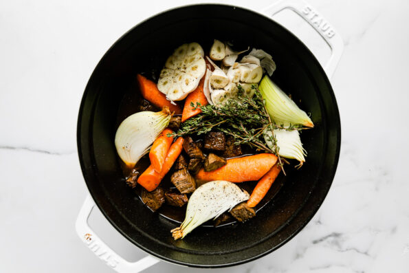 Browned beef, carrots, onion, garlic, and thyme fill a large white Staub Dutch oven. The pot sits atop a white and gray marble surface.