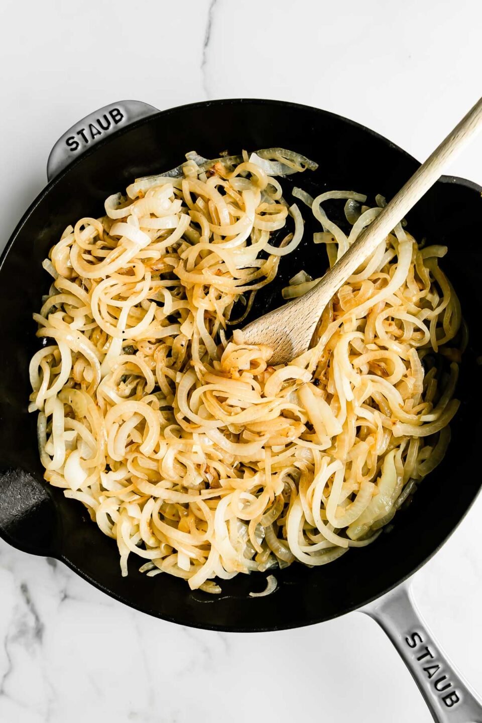 Thinly sliced sweet onions fill a large gray Staub cast iron skillet that sits atop a white and gray marble surface. The onions soften and wooden spoon rests inside of the skillet for stirring.