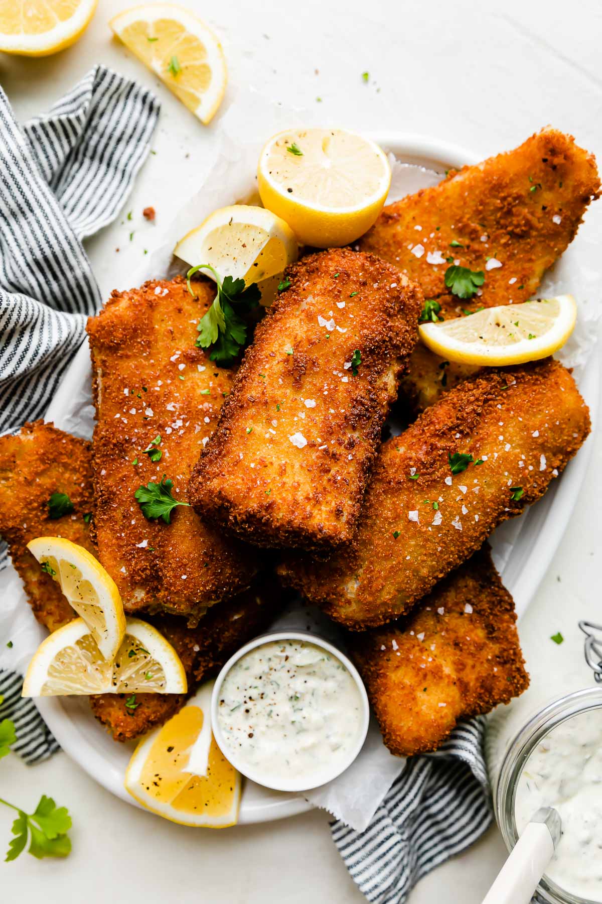 Fish fry pieces are arranged atop a white oval serving platter that sits a top a creamy white textured surface. The fish has been garnished with fresh parsley and flaky sea salt and is served alongside lemon wedges and a small bowl of tartar sauce. The platter is surrounded by a blue and white striped linen napkin, a small glass mason jar filled with tartar sauce with a spoon inside, fresh parsley leaves, and spent lemon wedges.