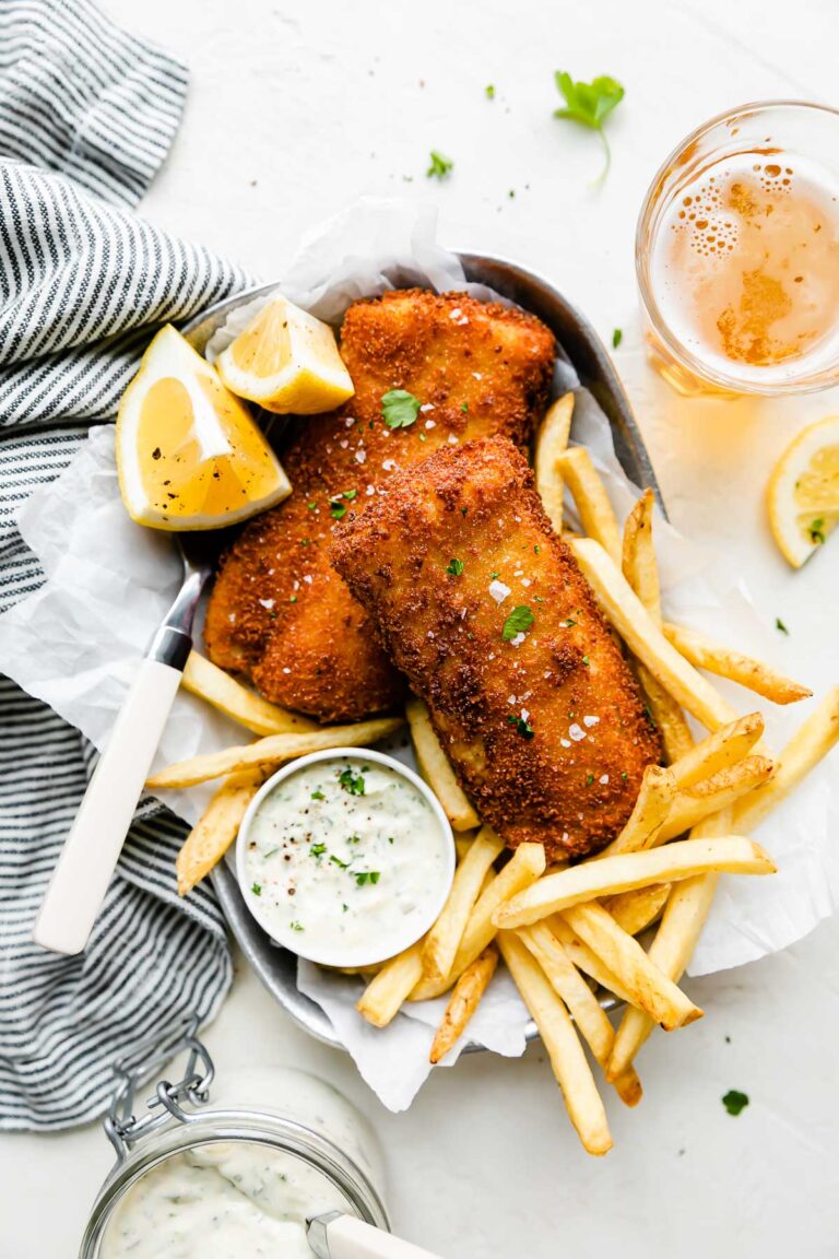 Fish fry pieces are arranged atop a metal oval tray that sits atop a creamy white textured surface. The fish has been garnished with fresh parsley and flaky sea salt and is served alongside lemon wedges, french fries, and a small bowl of tartar sauce. A fork rests on the tray and the tray is surrounded by a blue and white striped linen napkin, a small glass mason jar filled with tartar sauce with a spoon inside, loose fresh parsley leaves and lemon wedges, and a pint of beer.