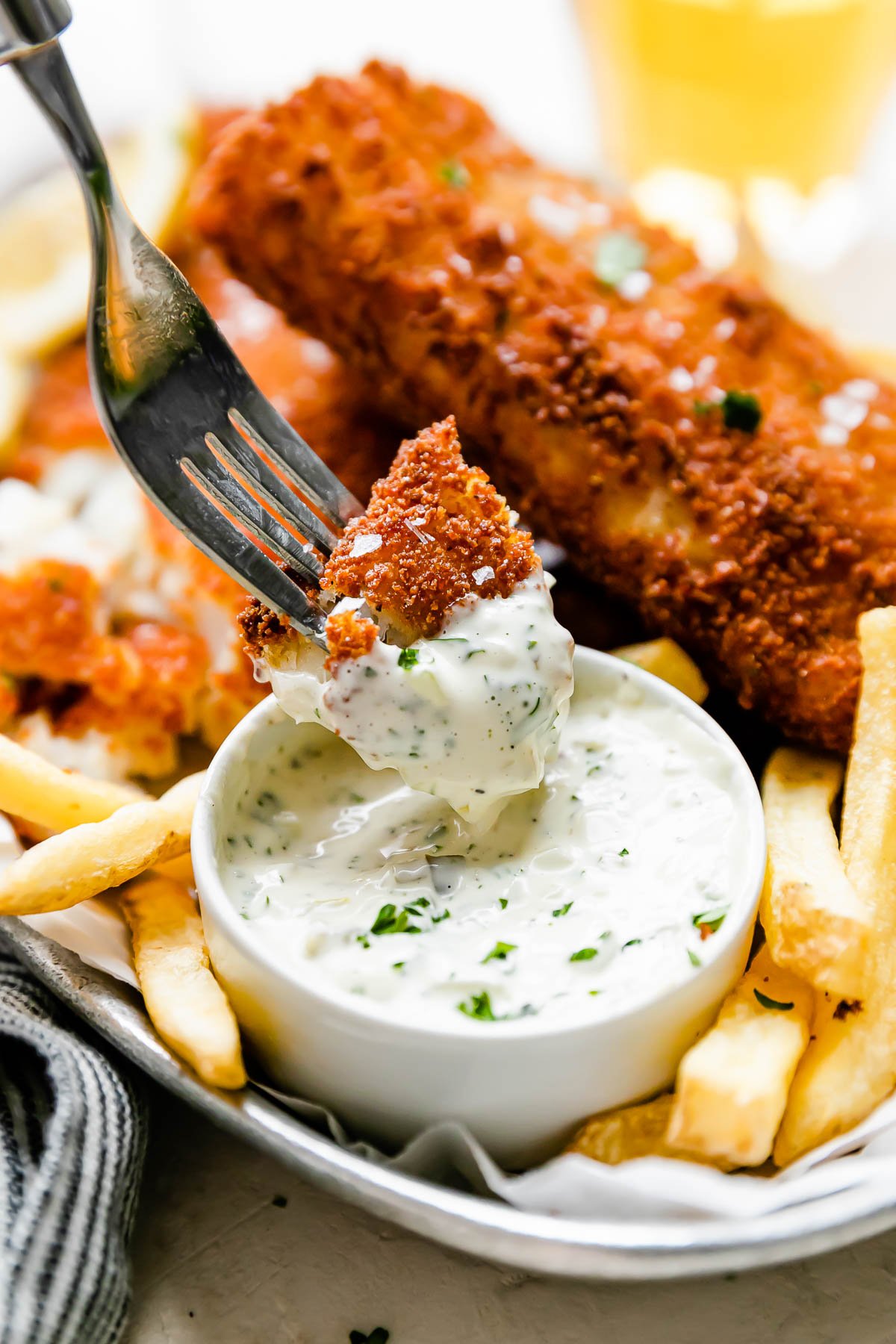 A side angle shot of fish fry pieces arranged atop a metal oval tray that sits atop a creamy white textured surface. The fish has been garnished with fresh parsley and flaky sea salt and is served alongside lemon wedges, french fries, and a small bowl of tartar sauce. A fork holds a piece of fried fish and dips it into the small white bowl filled with tartar sauce that sits atop the tray. A blue and white striped linen napkin rests alongside the tray.