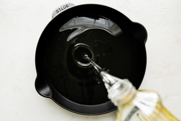 An overhead shot of frying oil being poured into a light gray Staub cast iron skillet that sits atop a creamy white textured surface.