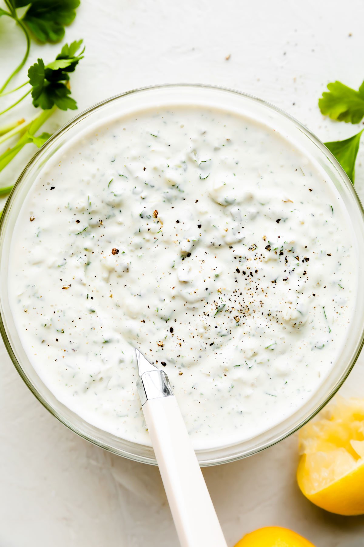 Mixed tartar sauce fills a small glass mixing bowl. A spoon with a white handle rests inside. The bowl sits atop a creamy white textured surface surrounded by fresh parsley and spent lemon halves.