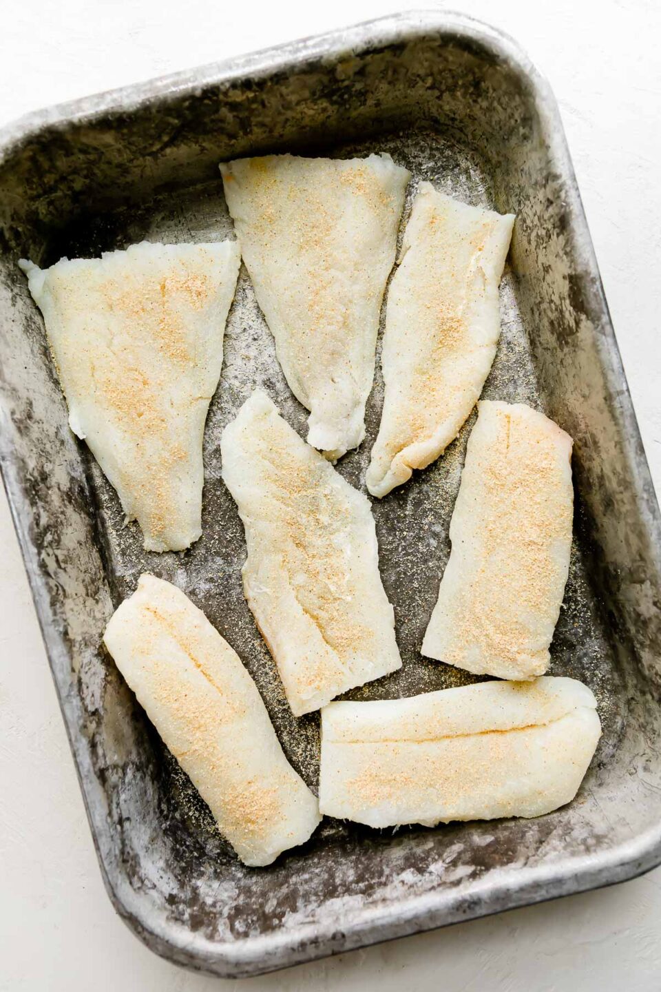 Prepared and seasoned cod fillets are arranged atop a metal baking sheet pan. The pan rests atop a creamy white textured surface.