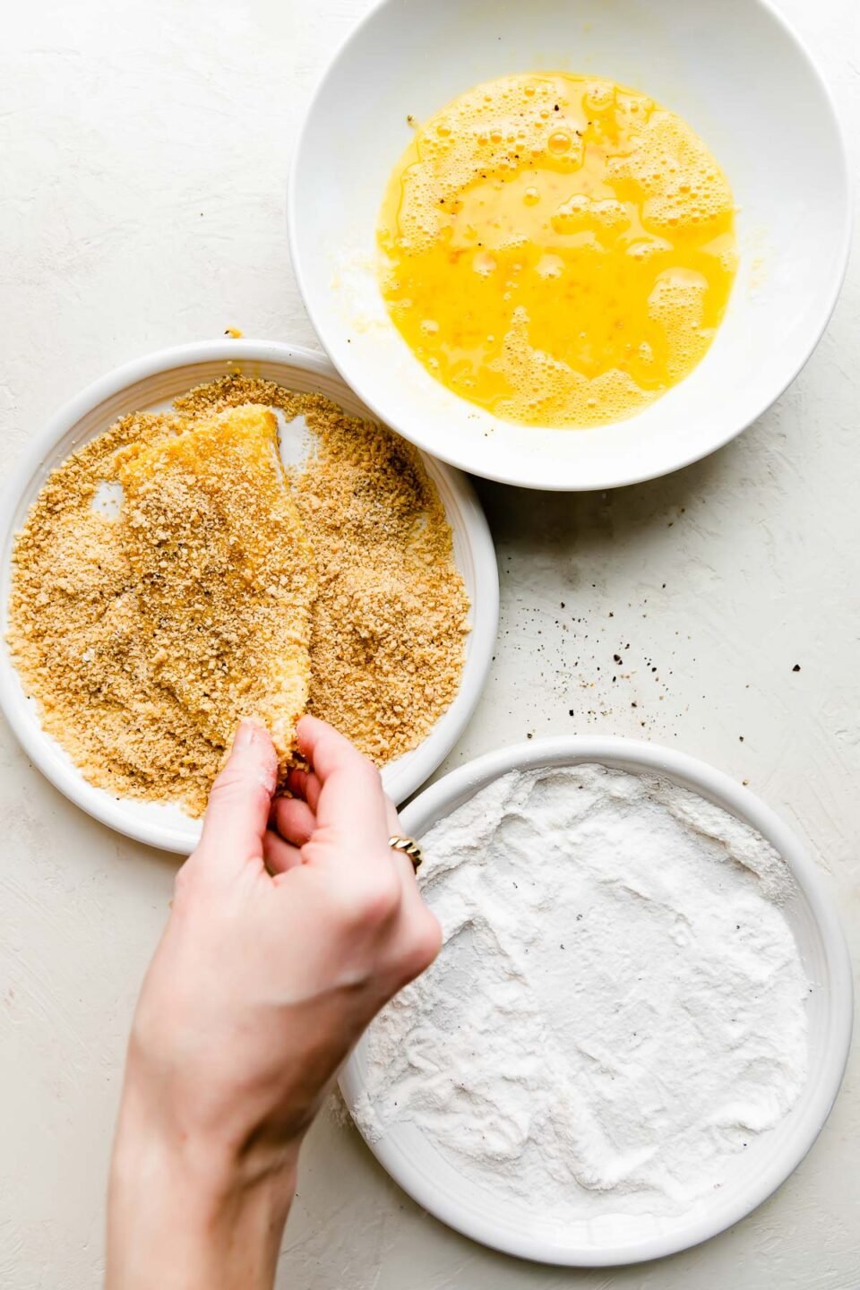 An overhead shot of a prepared dredging station for fish fry: a white plateful of rice flour, a second white plateful of panko breadcrumbs, and a third shallow bowl filled with beaten eggs. A woman's hand holds a cod fish filet as she coats the filet in panko breadcrumbs.